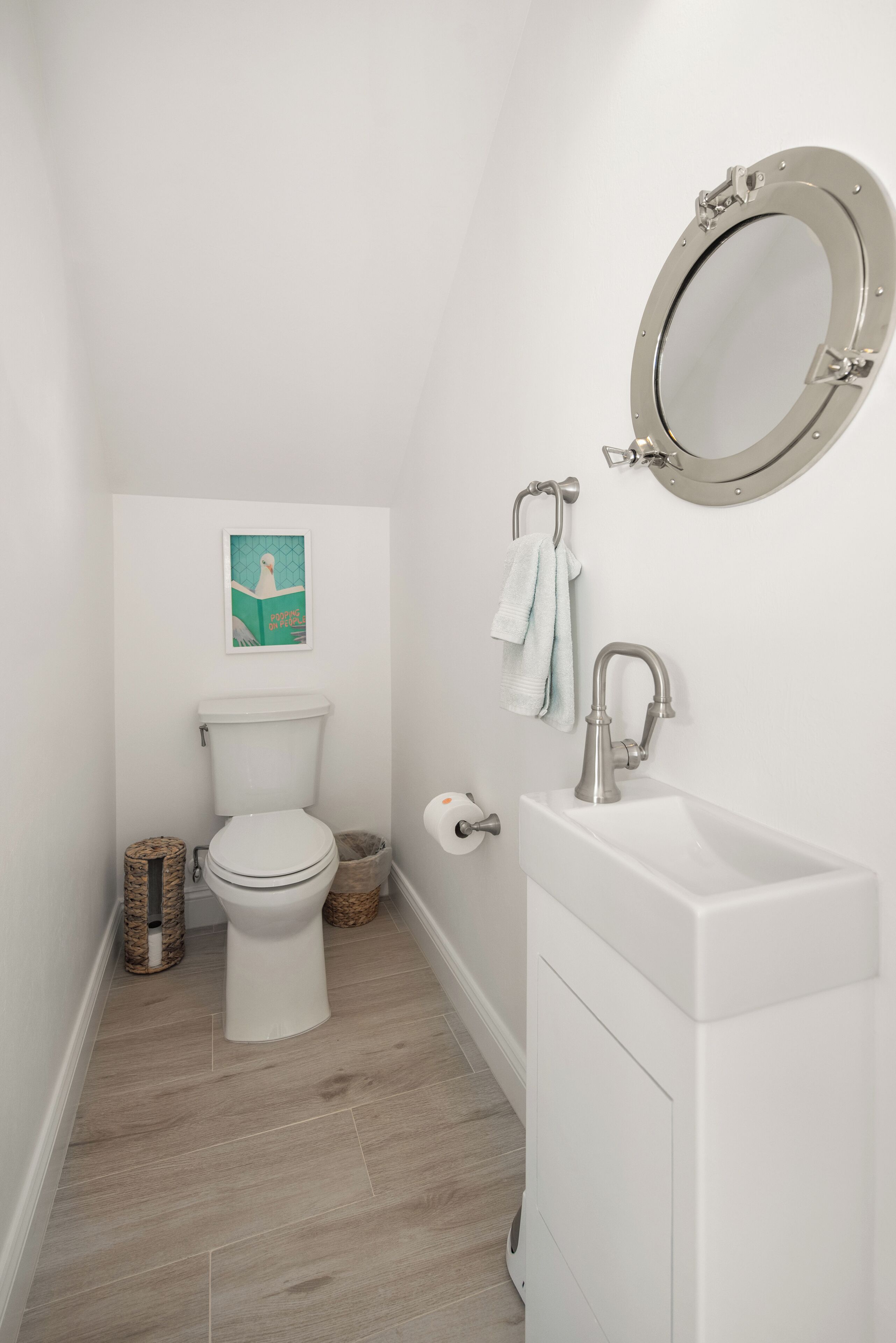 Un baño pequeño y blanco con inodoro, lavamanos y espejo. Hay un perchero y un cuadro en la pared.