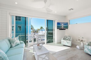 A spacious living room with a large sliding glass door leading to a balcony, a white sofa, a coffee table, and a television mounted on the wall. - On the beach cottage style cutie w/gorgeous beach views.  Walk to Times Square (Fort Myers Beach)