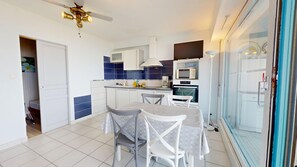 A compact kitchen with a dining area, white cabinets, blue tiles, and a ceiling fan. - Le Havre de Paix (Saint-Hilaire-de-Riez)