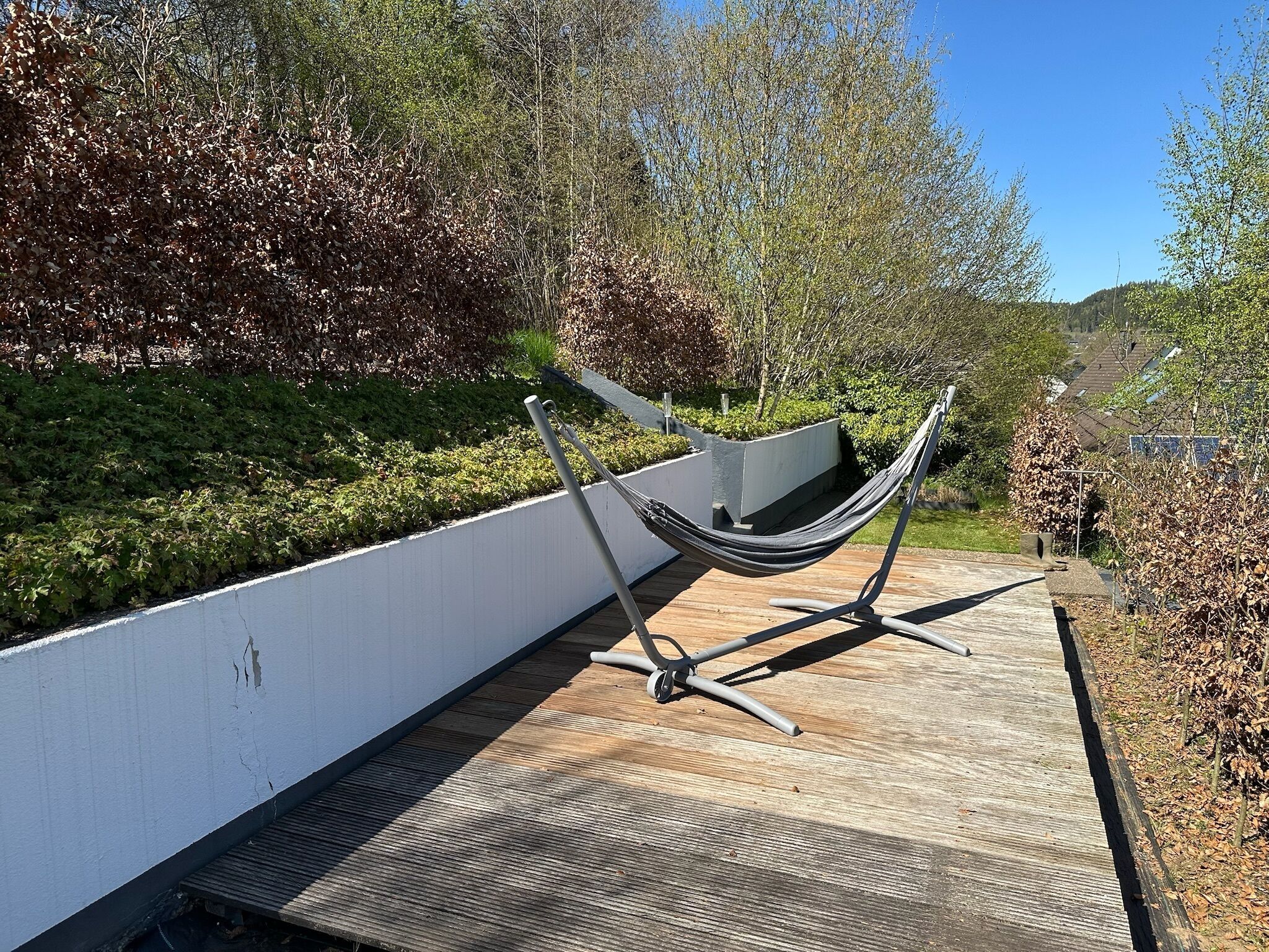 Een hangmat op een houten terras, met een witte muur en groene begroeiing op de achtergrond.