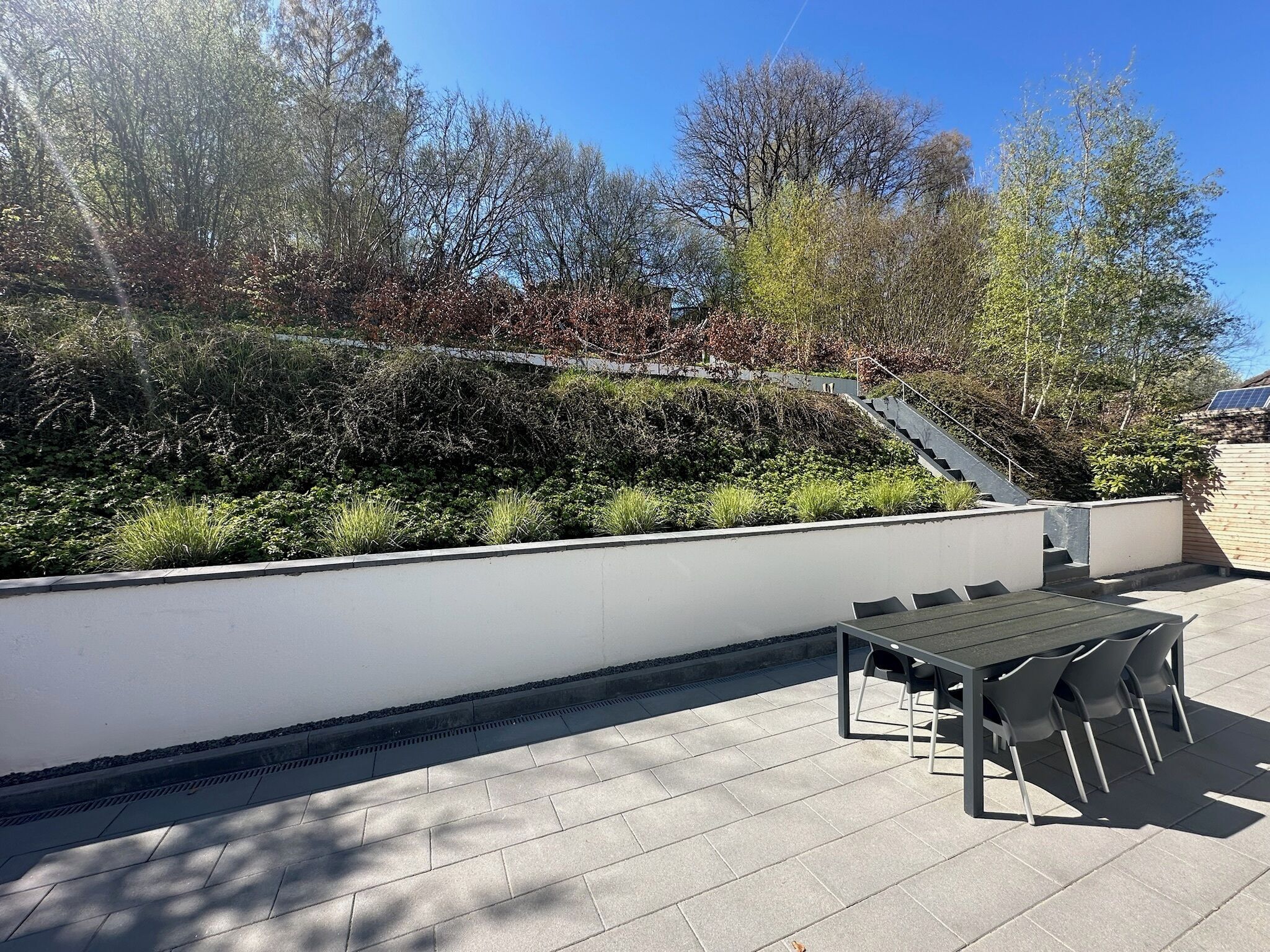 Eine Terrass mit Tisch und Stühlen, umgeben von einer niedrigen weißen Mauer und verschiedenen Pflanzen.