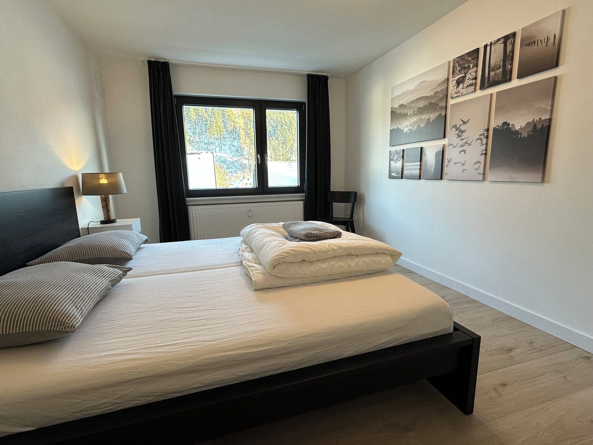 Un lit double avec une tête de lit sombre, deux tables de chevet avec des lampes, une fenêtre avec des rideaux et un mur orné de tableaux encadrés.