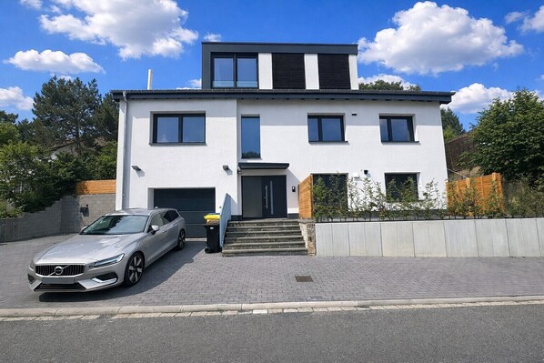 A modern two-story house with a garage, a driveway, and a car parked in front. - (NEW) Ferienwohnung Brachfeld – Space and relaxation in the Eifel National Park (Hellenthal)