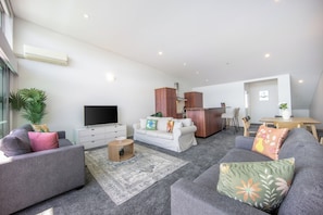 A modern living room with grey sofas, a flat-screen TV, a wooden cabinet, and a dining area with a table and chairs. - Harborfront Penthouse | Free Parking | Chill Vibes (Auckland)