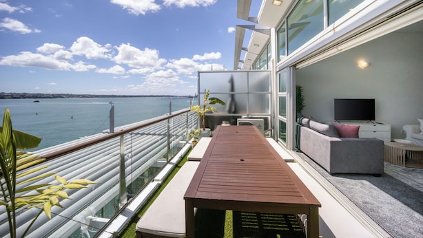 A modern balcony with a view of the sea, a wooden table, and a sofa with a TV. - Harborfront Penthouse | Free Parking | Chill Vibes (Auckland)