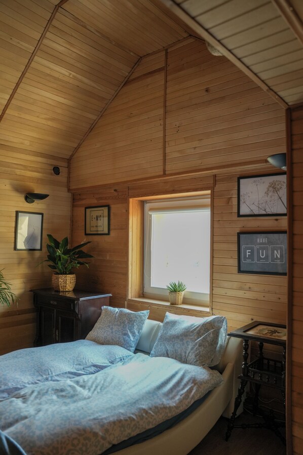 A cozy bedroom with a wooden ceiling, a large bed with patterned bedding, a window with a view, and a small table with a plant.