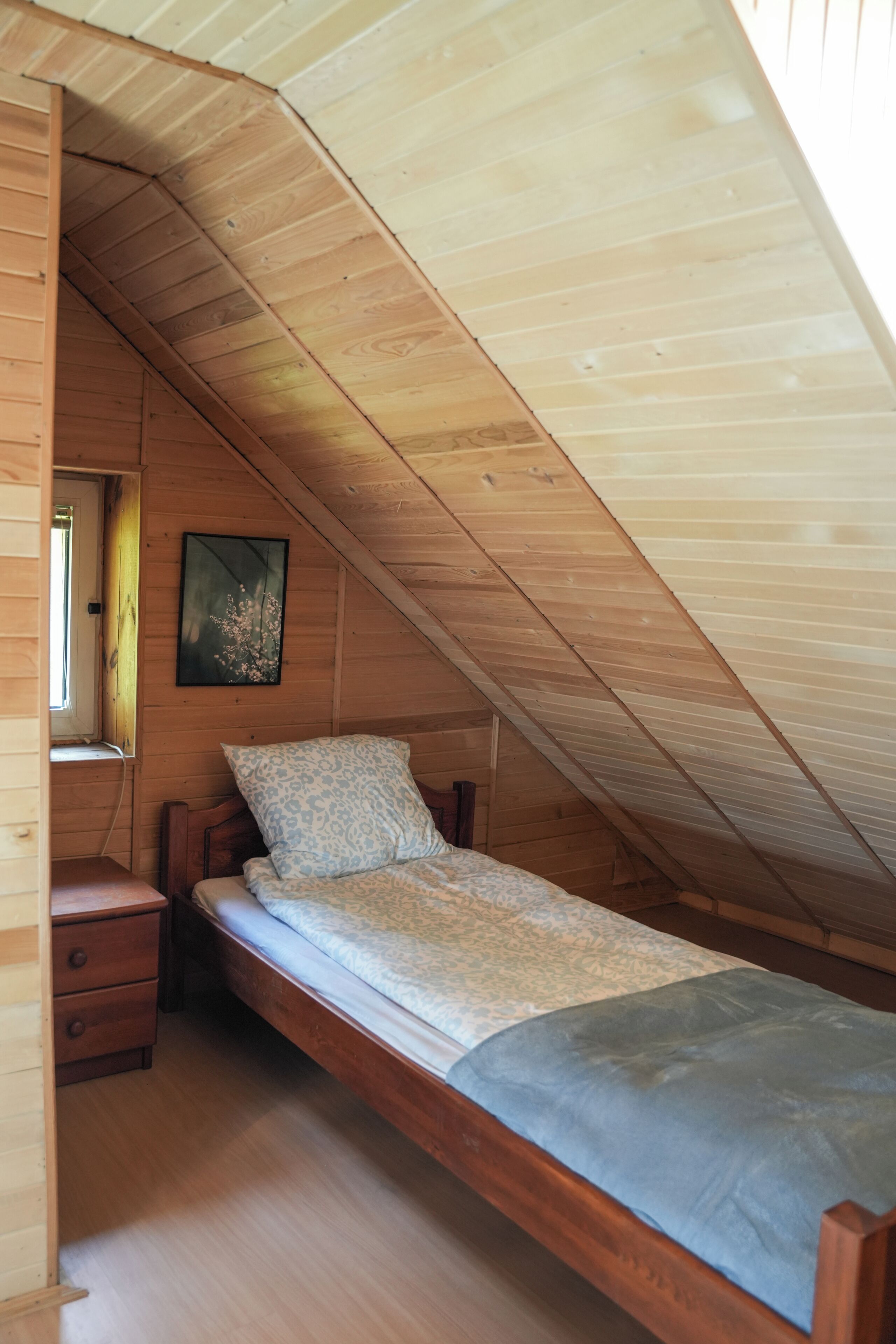 A small, wooden attic bedroom with a single bed, a nightstand, a window, and a framed picture on the wall.