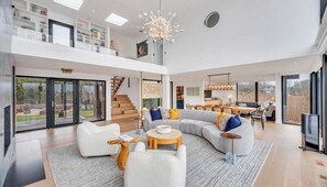 A spacious living area with a large sectional sofa, a coffee table, and a fireplace. There is a staircase leading to an upper level with a balcony. - Waterfront Southampton home with dock, pool, gym, great for groups (Southampton)