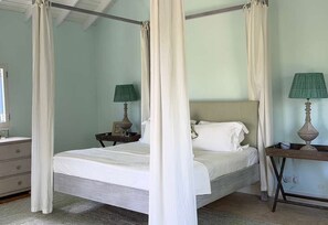 Una cama con dosel, ropa de cama blanca, dos mesitas de noche con lámparas y un ventanal con cortinas.