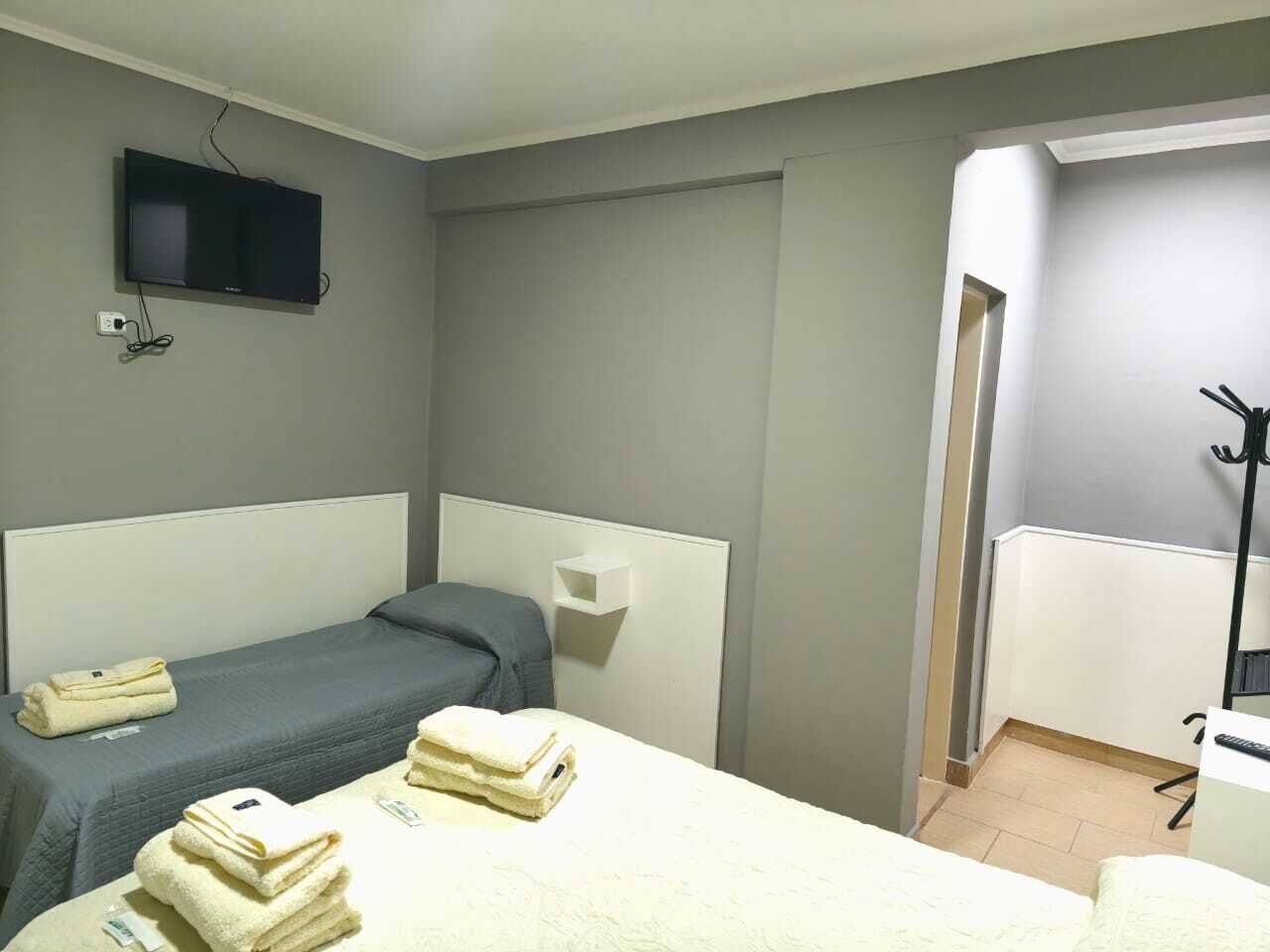 A hotel room with two single beds, a TV mounted on the wall, a small shelf, and a coat rack.