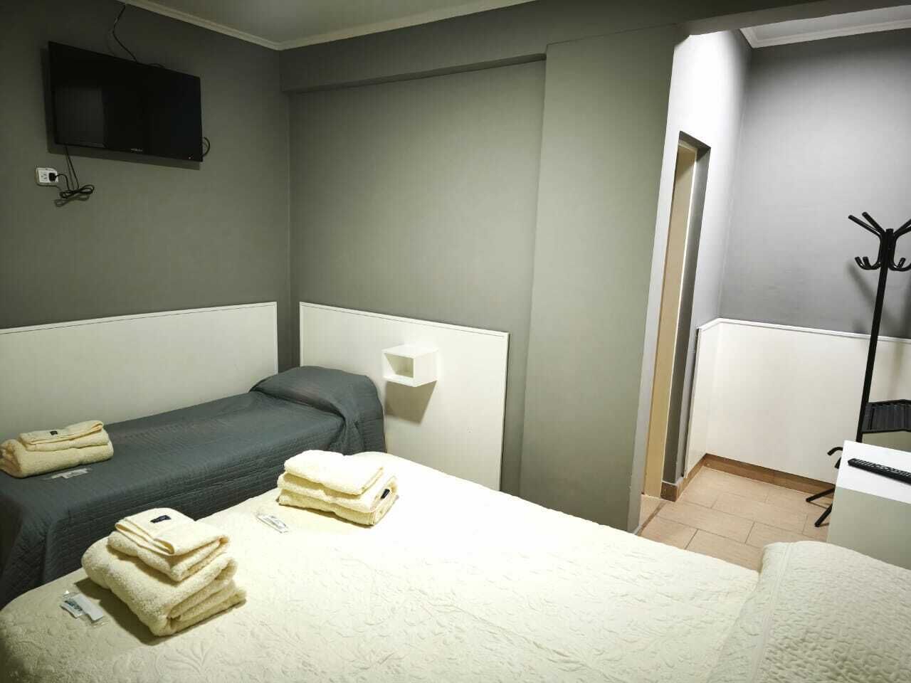 Habitación de hotel con dos camas individuales, un televisor montado en la pared, un perchero y un pequeño estante blanco.
