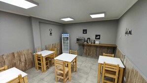 Une pièce comprenant six tables et chaises en bois, un coin cuisine équipé d’appareils électroménagers et une grande fenêtre laissant entrer la lumière naturelle.