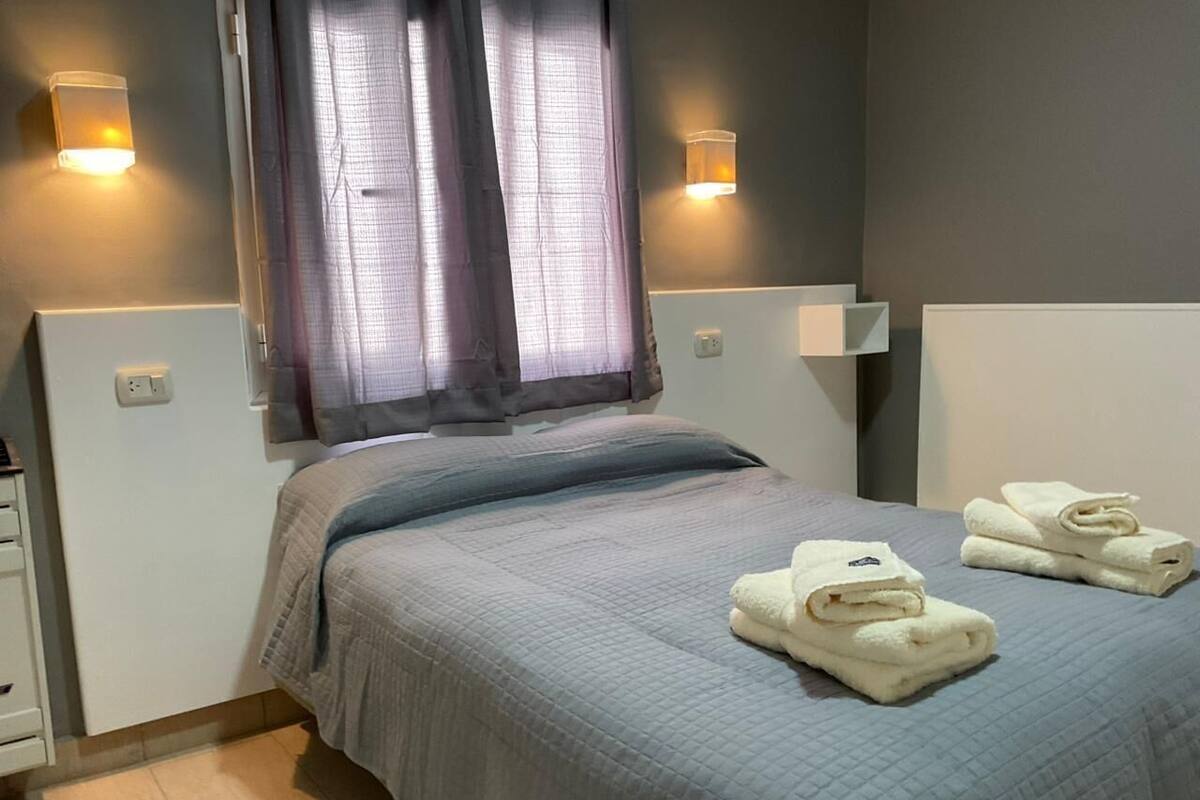 Un dormitorio con cama, dos lámparas de pared, una ventana con cortinas y dos toallas dobladas sobre la cama.