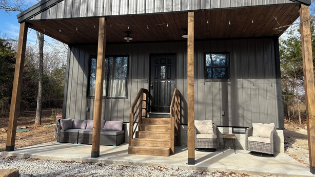 A rustic cabin with a covered porch, wooden steps, and wicker furniture. - Cozy new construction 2-bedroom house in serene Harrison with all amenities. (Harrison)