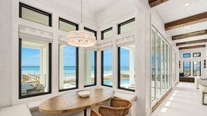 Amplia habitación con grandes ventanales que ofrecen vistas al océano, una mesa redonda de madera, sillas de mimbre y una lámpara moderna.