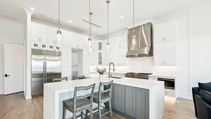 Eine moderne Küche mit weißen Schränken, einer großen Kücheninsel mit Barhocker, Edelstahlgeräten und Pendelleuchten.