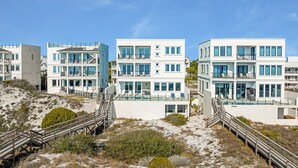 Une propriété située en bord de mer, composée de plusieurs bâtiments blancs, avec un escalier en bois menant à un espace piscine et un paysage sablonneux.