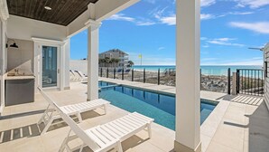 Un espace piscine avec des chaises longues, une vue sur la plage et un bâtiment surmonté d’un drapeau.