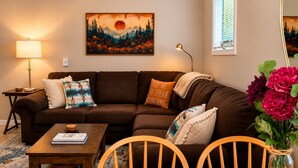 A living room with a brown sofa, a coffee table, a dining table with chairs, a painting on the wall, and a window with blinds. - Parkway Paradise Cozy Retreat Blue Ridge Parkway Family & Pet Friendly  (Laurel Springs)