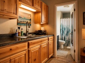 Une cuisine avec des meubles de rangement en bois, un évier et un plan de travail. Il y a un tableau au-dessus de l’évier et un rideau de douche orné d’un motif de forêt.