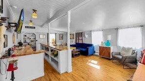 Een ruime woonkamer met een houten vloer, een blauwe bank, een grijze lederen fauteuil en een keuken met een houten aanrecht en een ballenautomaat.