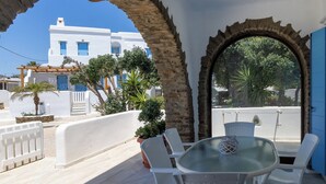 Un balcón con una mesa y sillas de vidrio, vista a un edificio blanco con puertas azules y un arco de piedra.