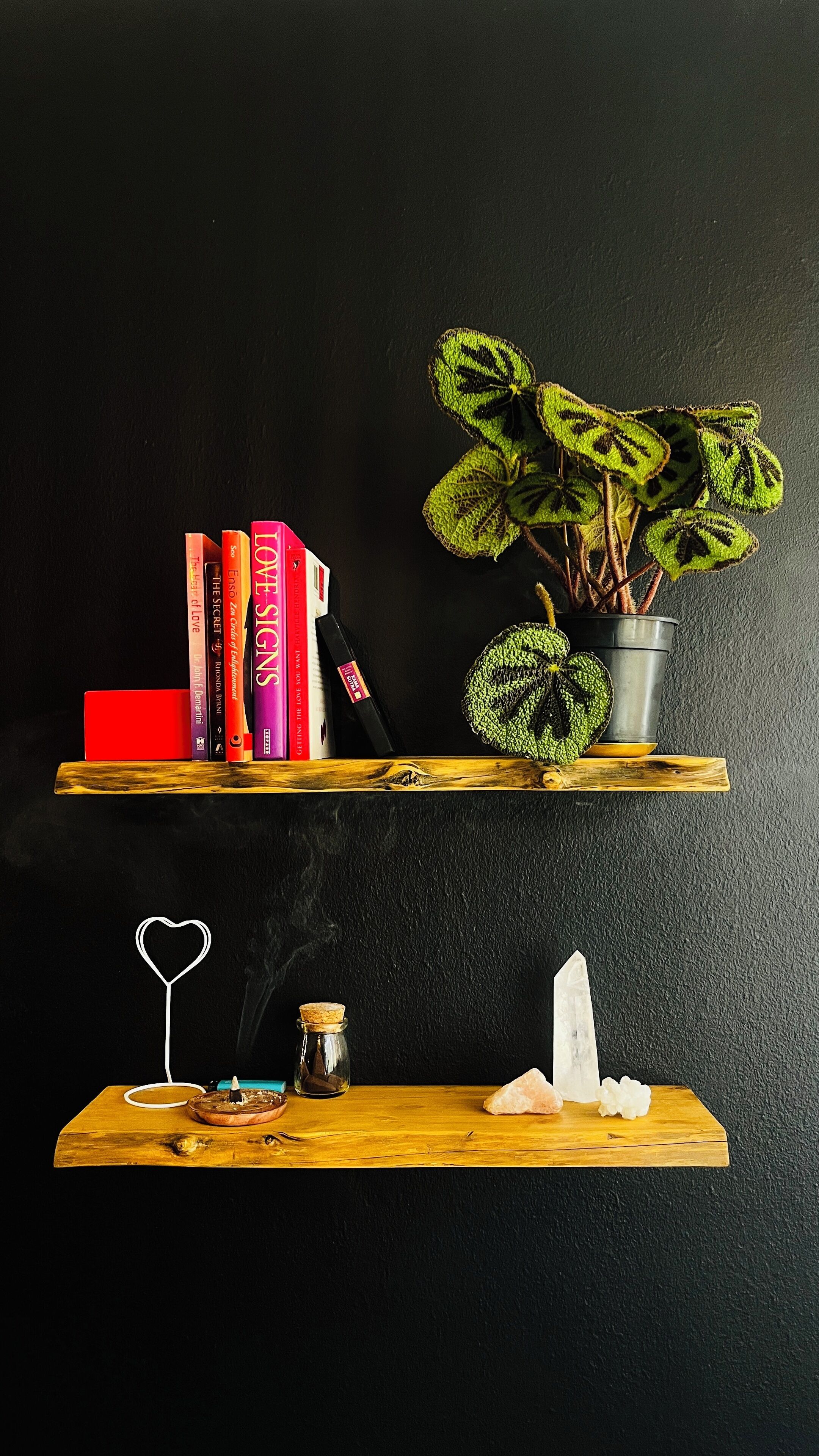 Two wooden shelves against a black wall, each holding various items. The top shelf has books, a small potted plant, and a red object. The bottom shelf has a heart-shaped candle holder, a small bottle, a crystal, and a pink stone.