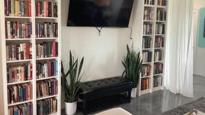 Un salón con un televisor de pantalla plana montado en la pared, rodeado de estanterías llenas de libros, dos plantas en macetas y un banco negro.