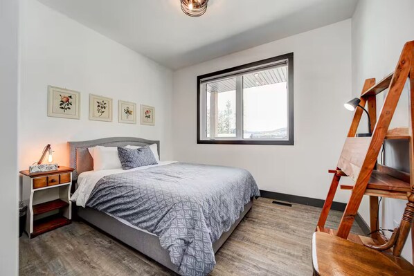 Ein Schlafzimmer mit Bett, Nachttisch, Lampe, einem Holzstuhl und einem Fenster mit Jalousien.