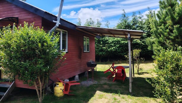 Une maison rouge avec un patio couvert, une cheminée et une cour avec des arbres et une chaise rouge.