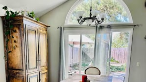Un comedor con una gran mesa de madera, bancos y una silla blanca. Se aprecia un aparador de madera con plantas encima, un candelabro y vistas a través de un ventanal amplio.