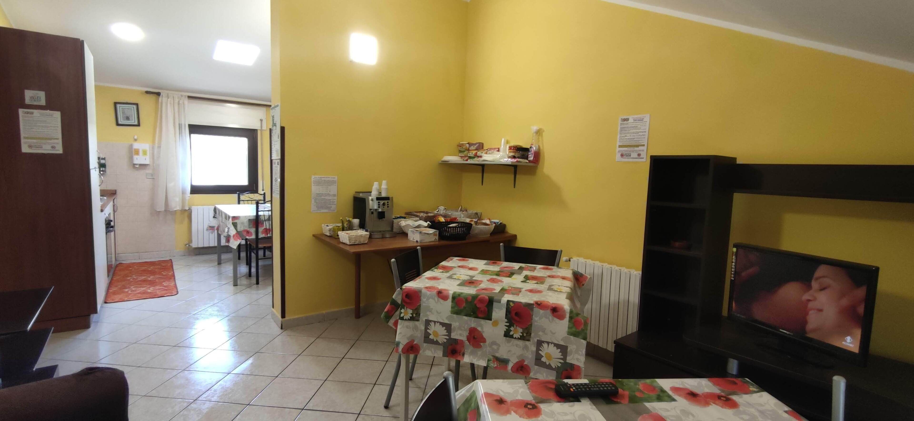 A room with a yellow wall, a dining table with a floral tablecloth, a television, and a small kitchen area with a microwave and a shelf with various items.
