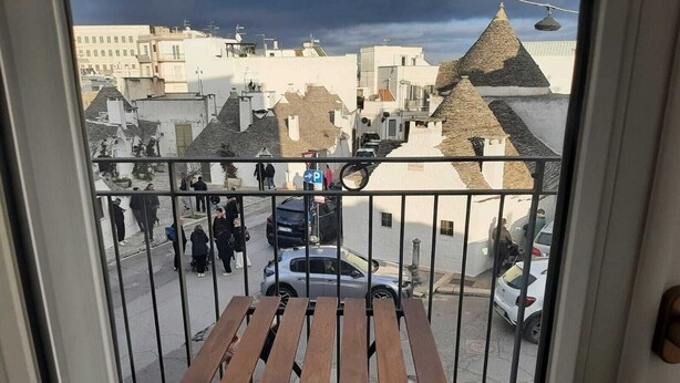 En balkon med et træbord og stole, der kigger ud over en gade med parkerede biler og mennesker.