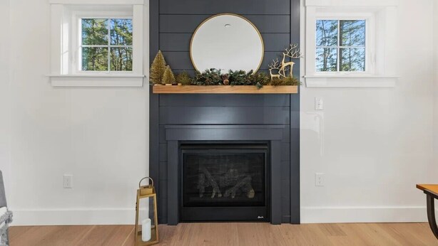 A modern living room with a dark fireplace, a round mirror above it, and two windows with a view of trees. - Beautiful new home near Portland in quiet Cumberland (Cumberland)