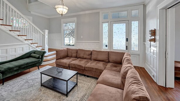 Uma sala de estar espaçosa com um amplo sofá de canto, uma poltrona verde e uma mesa de centro. Uma escada com guarda-corpos brancos é visível ao fundo.