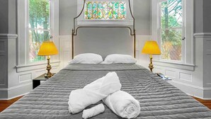 Cama bem arrumada com colcha cinza, duas luminárias de cabeceira e uma janela de vitral acima da cabeceira.