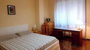 Un dormitorio con cama, escritorio, silla, lámpara, cómoda y un cuadro en la pared.