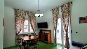 Un comedor con una mesa redonda de madera, sillas y un jarrón con flores. Hay un mueble de madera y un televisor de pantalla plana montado en la pared. La habitación tiene cortinas coloridas y una lámpara en el techo.