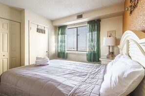 A neatly made bed with a white comforter and pillows, a bedside table with a lamp, and a window with green curtains.