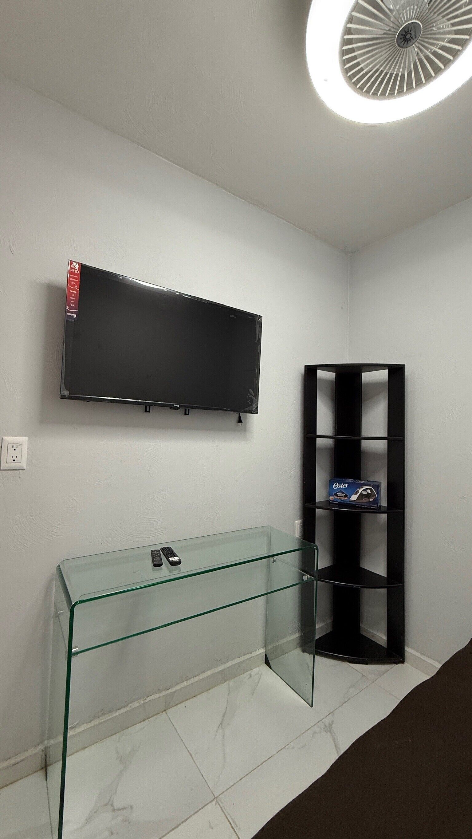 A room with a flat-screen TV mounted on the wall, a glass console table with a remote control, and a black shelving unit with a box labeled "Oster".