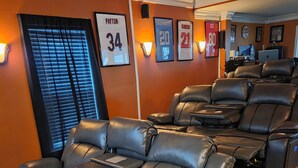 Sala de cinema com poltronas reclináveis de couro preto, uma tela grande e itens de coleção esportiva emoldurados na parede.