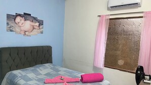A bedroom with a bed, a wall-mounted air conditioner, a window with a curtain, and a small table with a fan. - Maria Antônia's Family Home (Paraty)