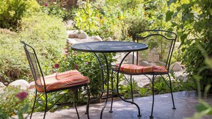A round metal table with two chairs, each with a cushioned seat and backrest. The setting is an outdoor garden with various plants and a wooden fence in the background. - Sunflower Hill Inn (Moab)