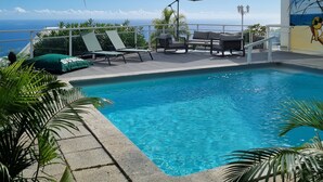 Ein Swimmingpool mit Liegestühlen und Sonnenschirmen, umgeben von Palmen und Blick aufs Meer.