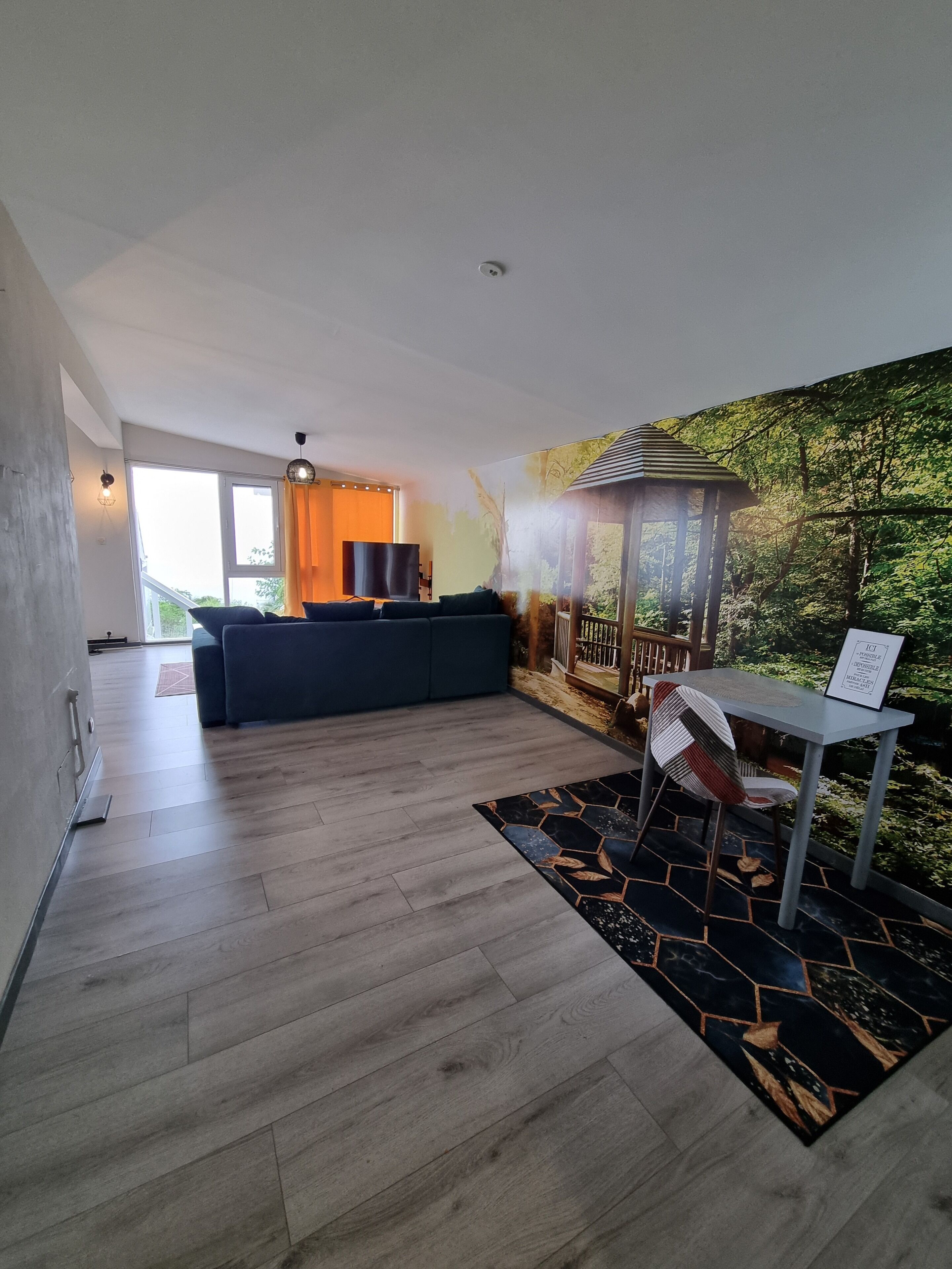 A modern living room with a large window, a dark sofa, a small table with a chair, and a wall mural of a forest.