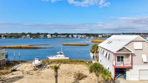 Une maison située sur la côte, dotée d’un balcon donnant sur une baie, avec des bateaux amarrés à proximité, et un ciel dégagé.