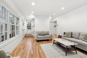 A spacious living room with hardwood flooring, a grey sofa, a white coffee table, and a large window with blinds. - Park Bound Bliss – Steps from Peninsula State Park! (fishcreek)
