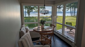 A sunroom with a view of a lake, a dining table with chairs, and a bench with cushions. - Lakefront Home with 200' of lakefront in the backyard (Northville)