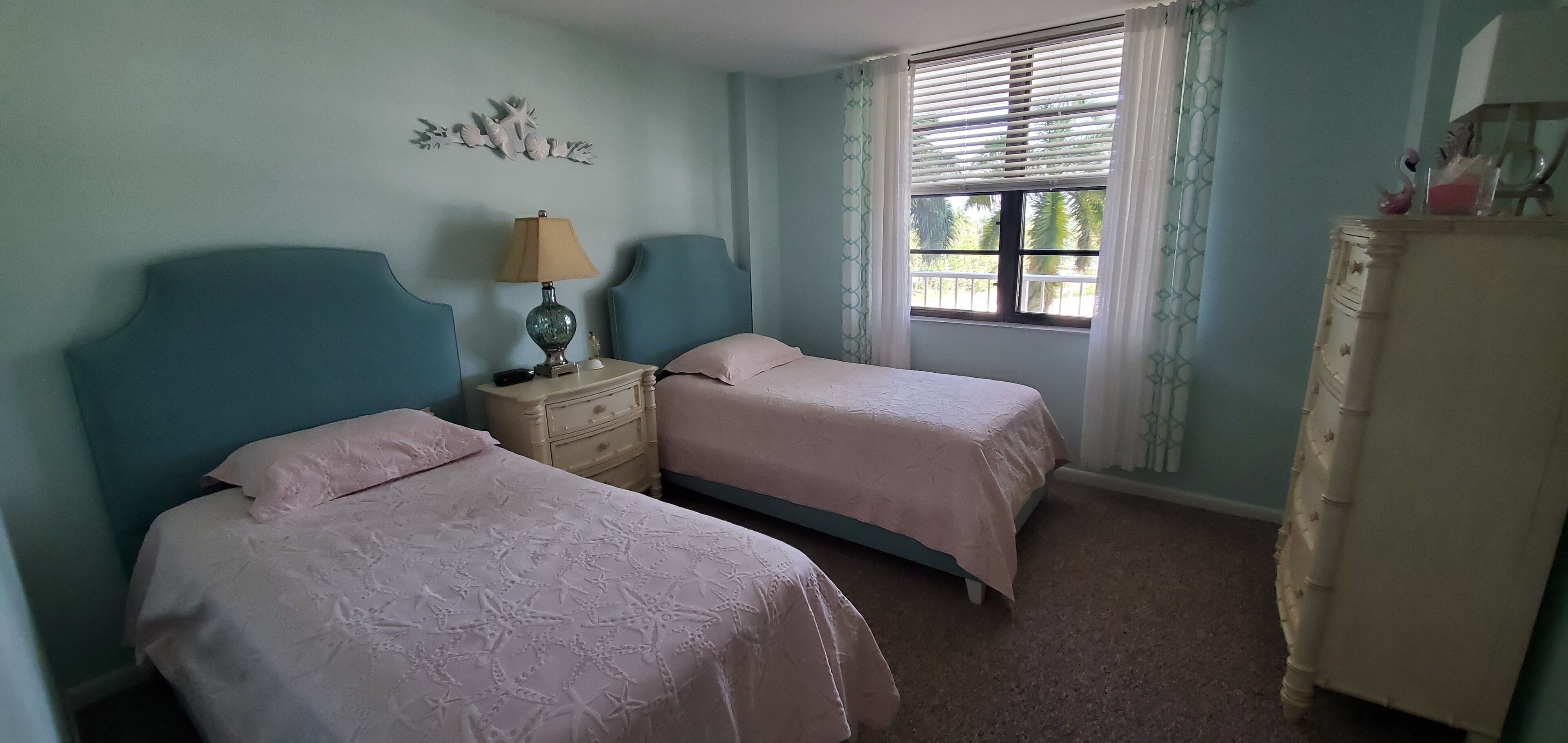 Een slaapkamer met twee bedden, een nachtkastje met een lamp, een dressoir en een raam met gordijnen.