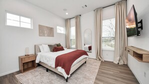 Ein modernes Schlafzimmer mit einem Bett, Nachttischen, einem an der Wand montierten Fernseher und einem großen Fenster mit Vorhängen.